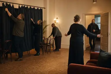 Voksne gør et kulturhus klar med stole og scenegardin, mens en vært tager imod en nyankommen ved den åbne dør.